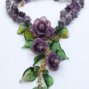 May include: A necklace featuring a strand of purple amethyst chip beads and a central pendant composed of three purple glass roses and several green glass leaves, all connected by a gold-toned metal chain and small gold-toned metal jump rings.