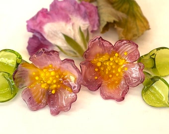 Könnte beinhalten: Zwei rosa Glasblumen mit gelben Zentren und grünen Glasblättern. Die Blumen haben eine strukturierte Oberfläche und sind durchscheinend.