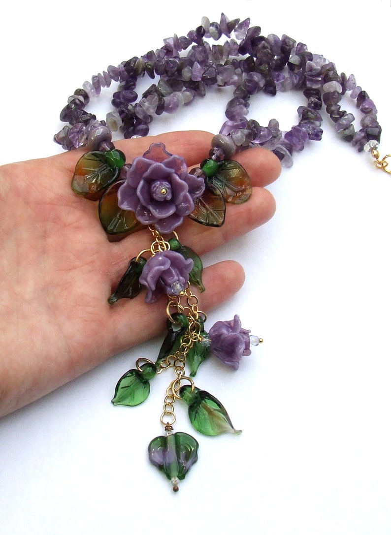May include: A necklace featuring a strand of purple amethyst chip beads and a central pendant arrangement of three carved purple glass roses and several green glass leaves, all connected by gold-toned metal links and small jump rings.