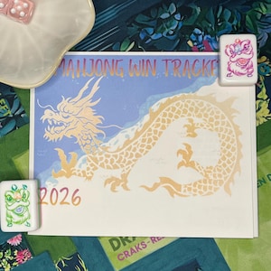 May include: A Mahjong win tracker card featuring a gold dragon illustration and the text 2026, accompanied by two pink dice in a white scalloped dish and two Mahjong tiles, one with a pink lion and one with a green lion graphic design.