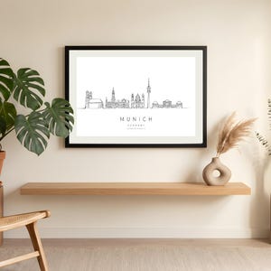 Pode incluir: Uma impressão artística com moldura preta apresentando um desenho de linha preta minimalista do horizonte de Munique, Alemanha, num fundo branco, com o texto MUNICH GERMANY impresso em letras maiúsculas pretas sob a ilustração.