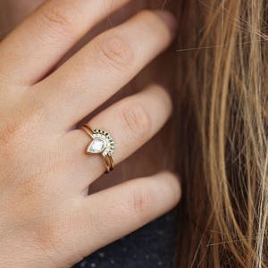 May include: A gold ring set with a pear-shaped diamond and a matching band with black diamonds. The ring is worn on the ring finger of a person's left hand.