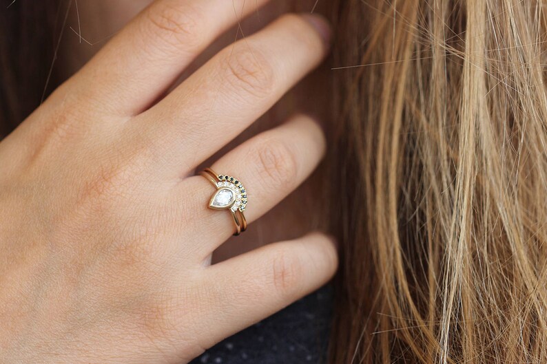 May include: A gold ring set with a pear-shaped diamond and a matching band with black diamonds. The ring is worn on the ring finger of a person's left hand.