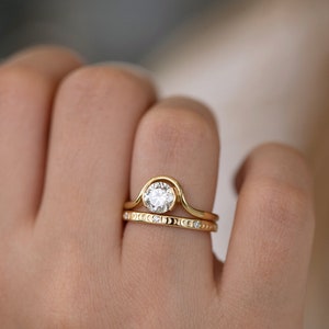 Puede incluir: Un anillo de compromiso de oro con un diamante redondo central y una banda curva. Una banda de oro a juego con peque&ntilde;os diamantes se lleva en el mismo dedo.