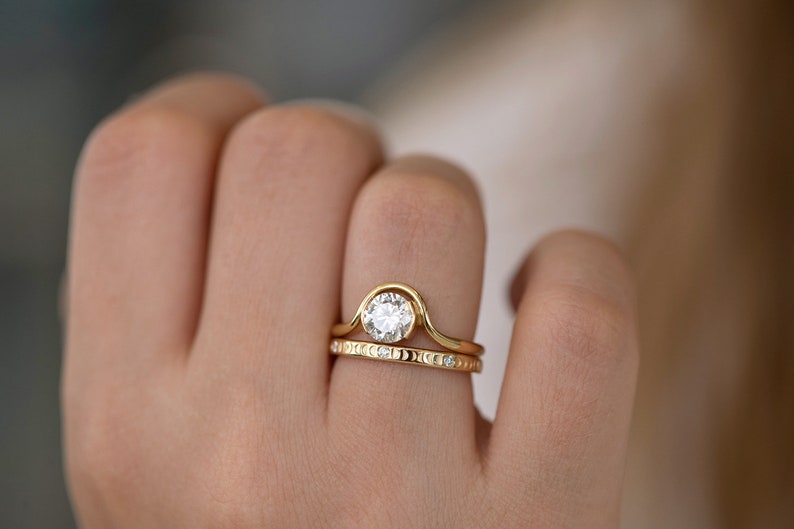 Puede incluir: Un anillo de compromiso de oro con un diamante redondo central y una banda curva. Una banda de oro a juego con peque&ntilde;os diamantes se lleva en el mismo dedo.