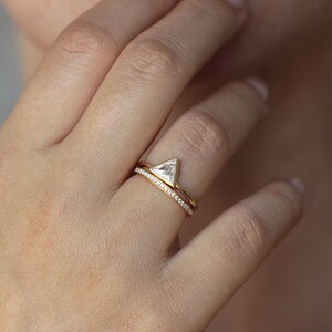 May include: A gold ring with a triangular diamond set in a prong setting. The ring is worn on the ring finger of a person's left hand. A thin gold band with small diamonds set in a pave setting is worn on the finger below the engagement ring.