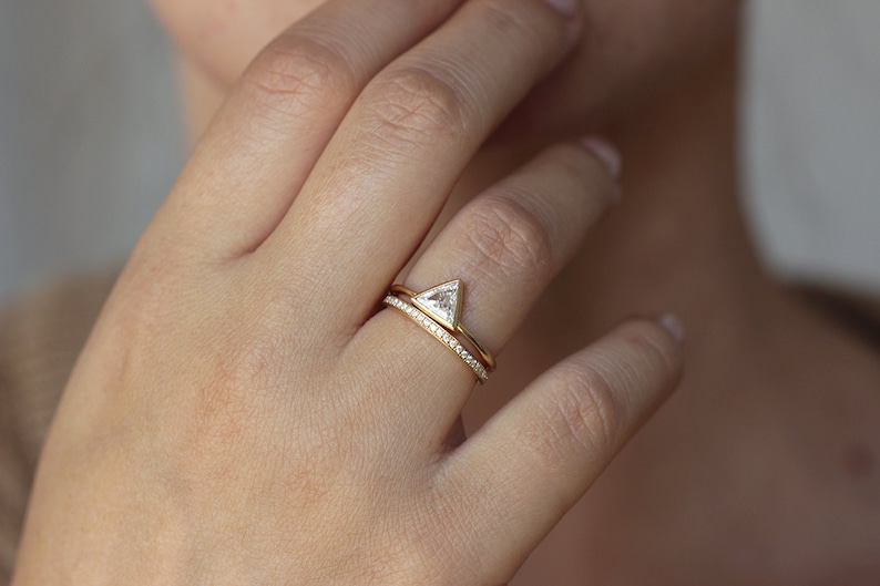 May include: A gold ring with a triangular diamond set in a prong setting. The ring is worn on the ring finger of a person's left hand. A thin gold band with small diamonds set in a pave setting is worn on the finger below the engagement ring.