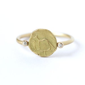 May include: A gold ring with a round coin-shaped center featuring an engraved design of a stylized animal. Two small diamonds are set on either side of the coin.