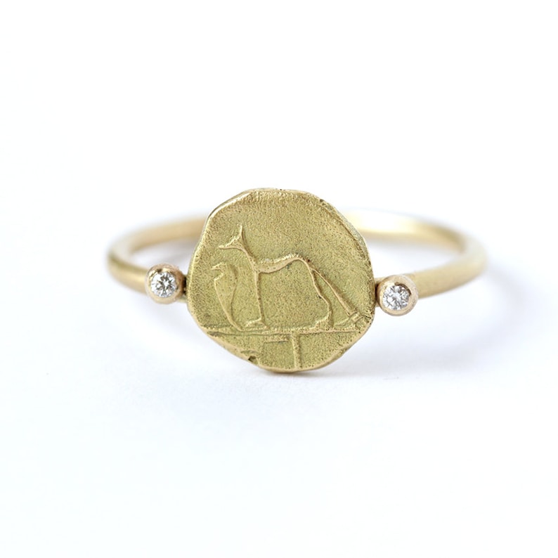 May include: A gold ring with a round coin-shaped center featuring an engraved design of a stylized animal. Two small diamonds are set on either side of the coin.