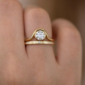 Puede incluir: Un anillo de oro engastado con un diamante redondo, engastado en bisel. El anillo de compromiso presenta una banda curva con un dise&ntilde;o de media luna. La banda de boda es una banda delgada con una fila de peque&ntilde;os diamantes y un dise&ntilde;o de media luna.