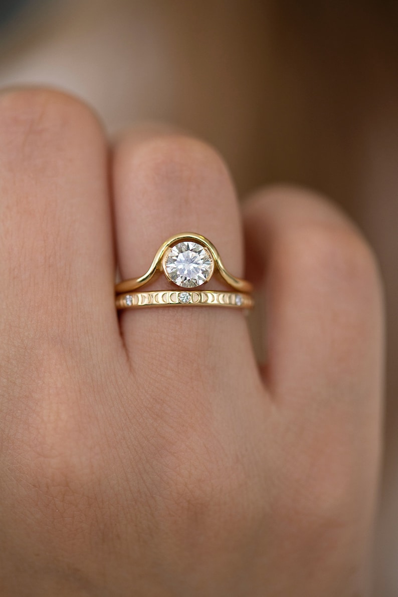 Puede incluir: Un anillo de oro engastado con un diamante redondo, engastado en bisel. El anillo de compromiso presenta una banda curva con un dise&ntilde;o de media luna. La banda de boda es una banda delgada con una fila de peque&ntilde;os diamantes y un dise&ntilde;o de media luna.