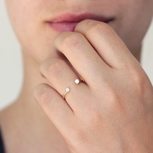 May include: A gold-colored ring with two small, clear gemstones set on either end of the open band. The ring is worn on a finger, with the gemstones positioned on the top and side of the finger. The ring is simple and elegant.