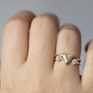 May include: A gold ring with a triangular diamond and two smaller round diamonds. The ring is on a person's finger.