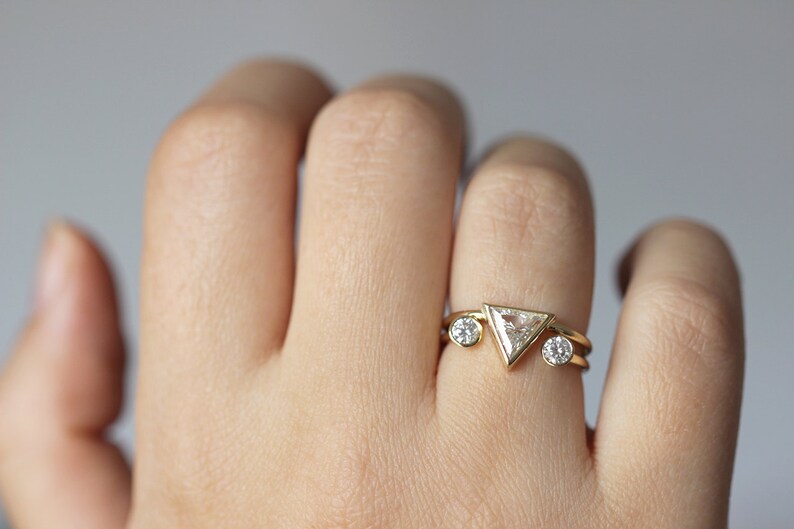 May include: A gold ring with a triangular diamond and two smaller round diamonds. The ring is on a person's finger.