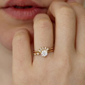 K&ouml;nnte beinhalten: Nahaufnahme einer Hand mit zwei goldenen Ringen. Der Hauptring hat einen gro&szlig;en, runden, klaren Edelstein, der von kleineren Steinen umgeben ist. Der zweite Ring ist ein schlichtes goldenes Band mit einem kleinen Stein.