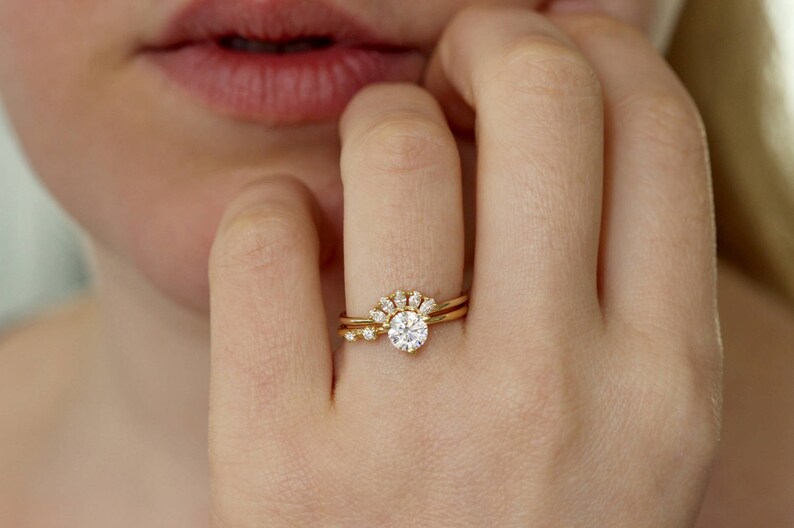K&ouml;nnte beinhalten: Nahaufnahme einer Hand mit zwei goldenen Ringen. Der Hauptring hat einen gro&szlig;en, runden, klaren Edelstein, der von kleineren Steinen umgeben ist. Der zweite Ring ist ein schlichtes goldenes Band mit einem kleinen Stein.