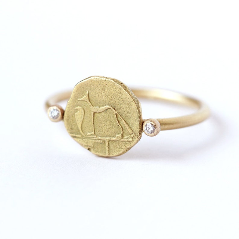 May include: A gold ring with a round coin-shaped design featuring a stylized animal figure. Two small diamonds are set on either side of the coin.