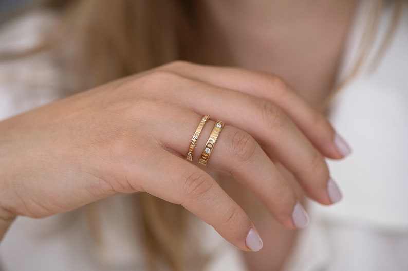 Puede incluir: Dos anillos de oro con peque&ntilde;os diamantes en un dedo. El anillo de abajo tiene una banda texturizada y el anillo de arriba tiene una banda lisa.