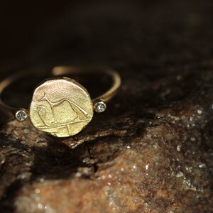 May include: A gold ring with a round, engraved coin-shaped design featuring a stylized animal figure. The ring has two small, clear diamonds on the band.