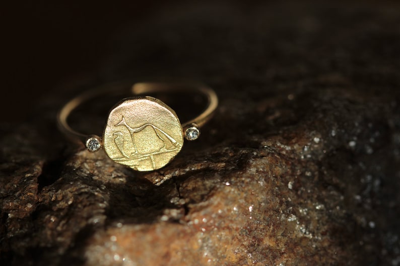 May include: A gold ring with a round, engraved coin-shaped design featuring a stylized animal figure. The ring has two small, clear diamonds on the band.