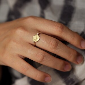 May include: A gold ring with a round, textured, coin-shaped design and two small, clear gemstones on the band.