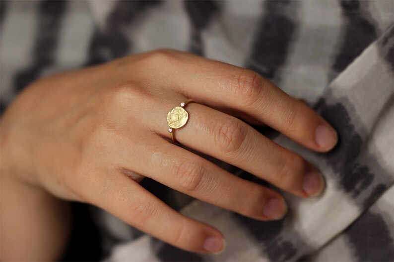 May include: A gold ring with a round, textured, coin-shaped design and two small, clear gemstones on the band.