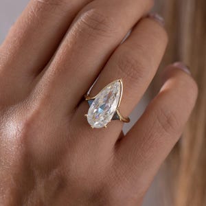 May include: A gold ring featuring a large, pear-shaped, clear gemstone. The ring has a delicate band and small, dark accents on either side of the gemstone. The ring is set against a blurred background.