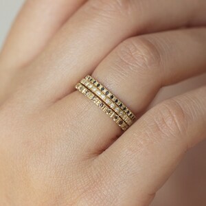 May include: Three gold stacking rings with pave diamonds and black diamonds. The rings are worn on a finger.