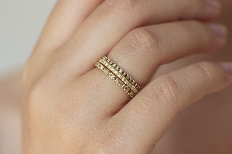 May include: Three gold stacking rings with pave diamonds and black diamonds. The rings are worn on a finger.