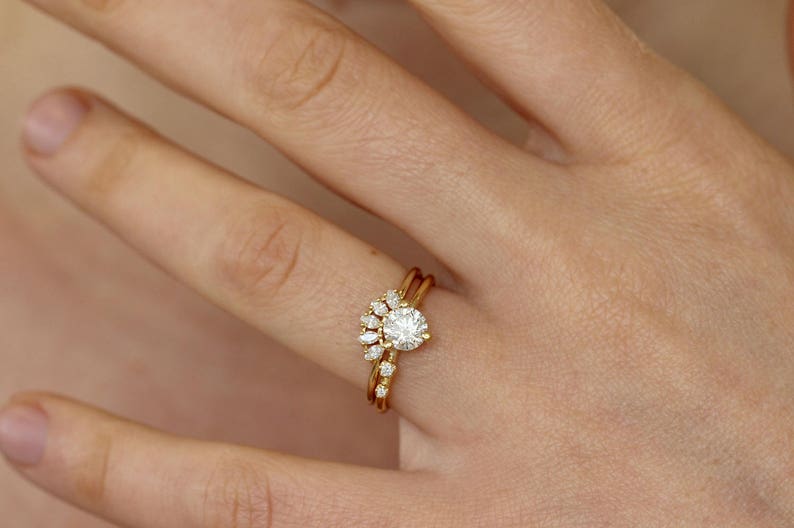 K&ouml;nnte beinhalten: Ein goldener Ringsatz mit einem runden, klaren Edelstein als Mittelpunkt, erg&auml;nzt durch kleinere, klare Edelsteine in f&auml;cherf&ouml;rmiger Anordnung. Das Set enth&auml;lt ein gebogenes Band mit zus&auml;tzlichen kleinen Edelsteinen.