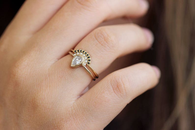 May include: A pear-shaped diamond engagement ring with a delicate band, paired with a matching band featuring a row of black diamonds.