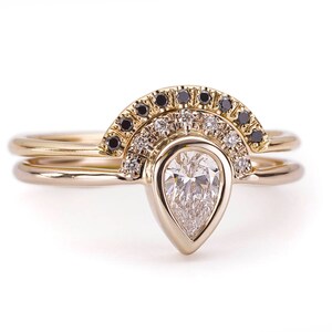 May include: A pear-shaped diamond engagement ring set with a delicate band featuring a row of black diamonds and a row of white diamonds. The ring is set in yellow gold.