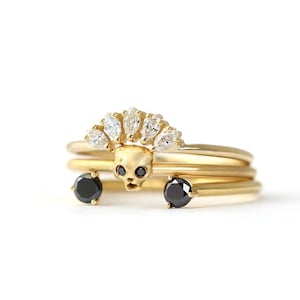 K&ouml;nnte beinhalten: Ein Satz goldener Ringe. Der zentrale Ring hat ein Totenkopf-Design mit schwarzen Edelsteinen f&uuml;r die Augen. Zwei schwarze Edelsteine flankieren den Sch&auml;del. Der obere Ring hat eine Reihe klarer Edelsteine. Vor wei&szlig;em Hintergrund.