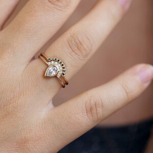 May include: A pear-shaped diamond engagement ring with a delicate band, paired with a matching wedding band featuring a row of black diamonds set in a curved shape.