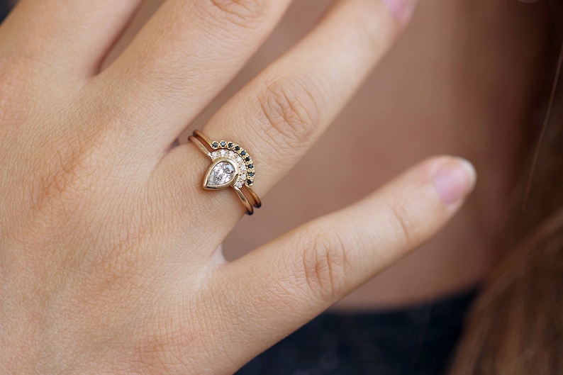 May include: A pear-shaped diamond engagement ring with a delicate band, paired with a matching wedding band featuring a row of black diamonds set in a curved shape.