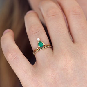 May include: A gold ring set with a pear-shaped emerald gemstone and a small diamond. The ring is paired with a thin band set with black diamonds.