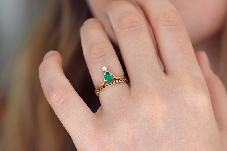 May include: A gold ring set with a pear-shaped emerald gemstone and a small diamond. The ring is paired with a thin band set with black diamonds.
