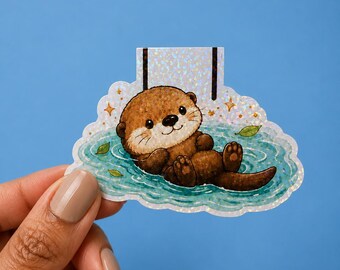 May include: A magnetic bookmark featuring a brown otter floating on its back in blue water. The design includes small green leaves and gold star accents on a holographic background that shimmers with light. The otter has a smiling face and paws.