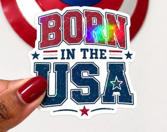 May include: A sticker with the text "BORN IN THE USA" in red and blue, with a holographic effect. The sticker is held in front of a Captain America shield.