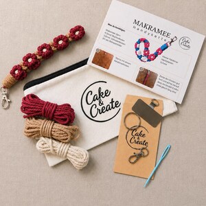 Könnte beinhalten: Ein Makramee-Bastelset mit einem fertigen rot-beigen Blumenarmband, einem weißen Stoffbeutel mit der Aufschrift Cake & Create, drei Garnbündeln in Rot, Beige und Creme, einem Metallring, einem Karabiner, einer Nadel und einer Anleitung.