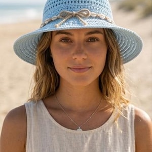 May include: A light blue crochet sun hat featuring a wide brim and a decorative band made of natural twine. The band is adorned with small white seashells and a central bow detail. The hat has a textured, open-weave pattern throughout the crown.