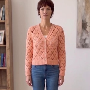 May include: A peach-colored, open-weave crochet cardigan with a V-neck and button closure. The long-sleeved sweater is paired with blue jeans. The cardigan's intricate pattern and soft color create a delicate, feminine aesthetic.