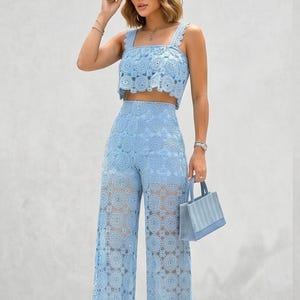 May include: A light blue lace two-piece set, featuring a crop top with scalloped edges and wide-leg pants. The outfit is paired with a matching handbag. The set is made of a delicate, open-weave fabric.