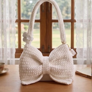 Pode incluir: Uma bolsa branca de crochê em forma de laço, com uma alça combinando e um pequeno detalhe floral. A bolsa tem uma aparência texturizada e uma base retangular. Um pequeno pingente pende da alça.