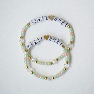 Pode incluir: Duas pulseiras de contas com esferas redondas em verde claro e rosa suave, intercaladas com esferas douradas metálicas e cristais transparentes facetados. Cada pulseira inclui contas brancas com letras que formam PIXIE DUST e um coração dourado.