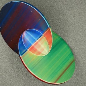 May include: Two overlapping oval-shaped objects with a smooth, iridescent finish. The top oval features deep blue, purple, and red tones, while the bottom oval displays vibrant green, orange, and red hues, creating a layered, multi-colored effect.