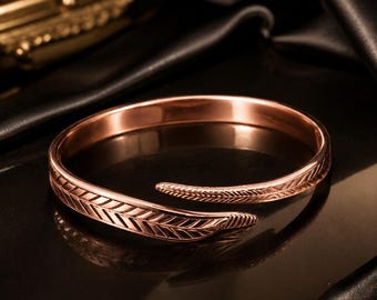 May include: A rose gold-toned open cuff bracelet featuring a textured feather design that wraps around the wrist. The metal surface has a polished finish with engraved details along the band, creating a continuous leaf-like pattern on both ends.