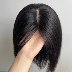 Può includere: Un posticcio di capelli castano scuro con una riga dall'aspetto naturale. I capelli sono lisci e sembrano essere capelli umani. Il posticcio è tenuto in una mano, mostrando il frontale in pizzo e la consistenza dei capelli. Progettato per aggiungere volume.