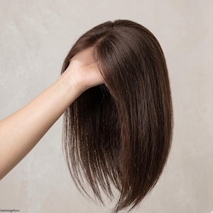 May include: A brown wig with long, straight hair. The wig is held by a hand, showcasing the part and the natural-looking strands. The hair appears to be a rich, dark brown color.
