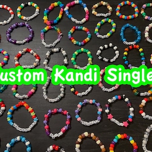 Può includere: Una collezione di braccialetti Kandi colorati, ognuno con una combinazione unica di perline e dettagli con lettere. I braccialetti sono in vari colori, tra cui rosso, blu, verde e viola. Il testo "Custom Kandi Singles!" è visualizzato in verde.